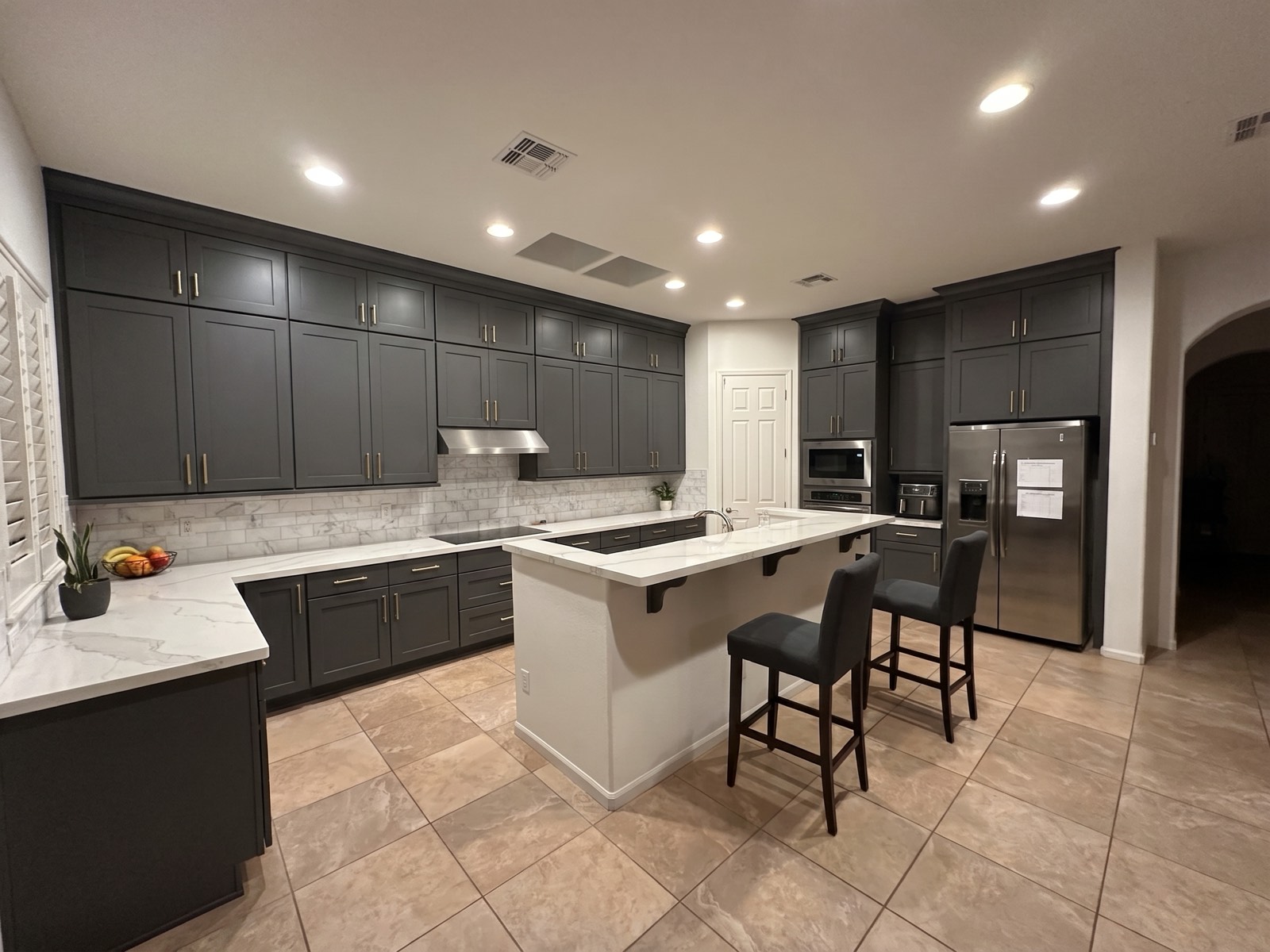 The same kitchen, AI-rendered: charcoal flat-panel shaker cabinets with slim
                  brass pulls, white marble-look quartz counters, a white marble subway backsplash,
                  and cleared worktops. The room, the island, the stools and the floor are unchanged.