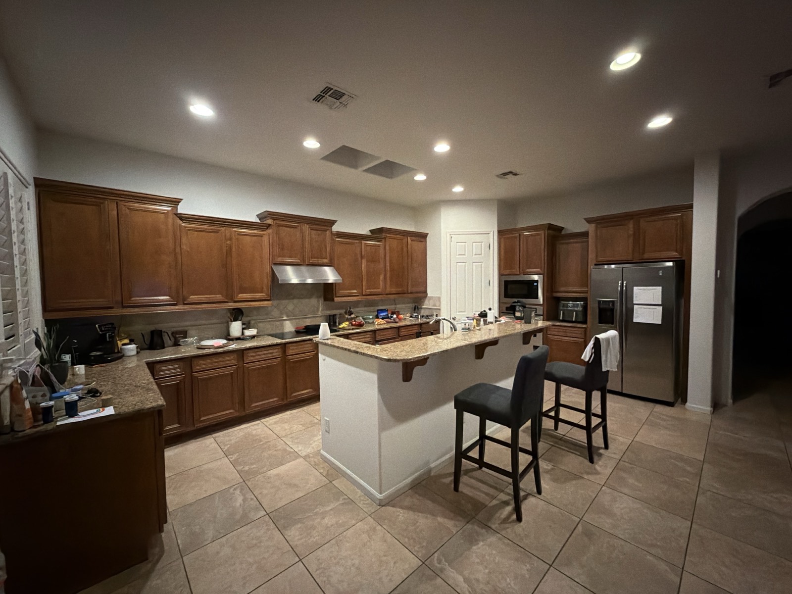 A lived-in Scottsdale kitchen photographed on a phone: dark maple raised-panel
                  cabinets, speckled granite counters, a peninsula with two bar stools, and small
                  appliances and papers spread across the worktops.